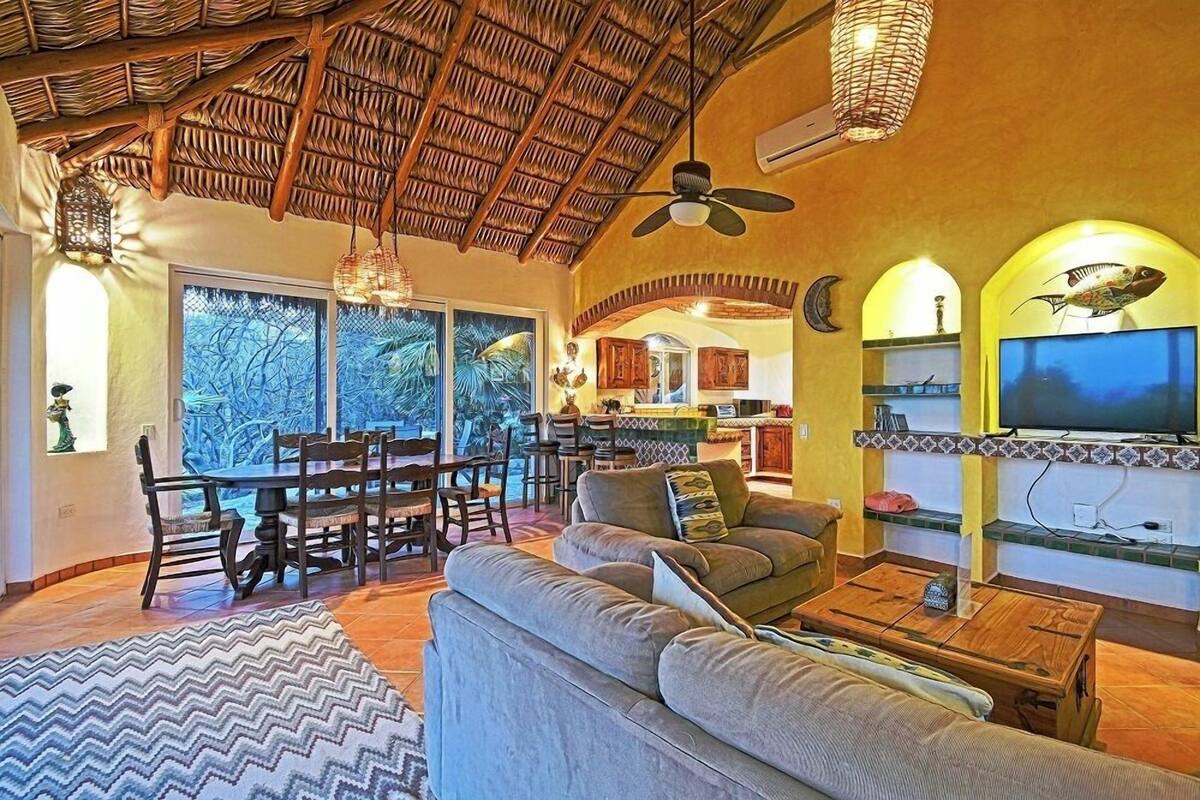 A spacious living area with a thatched ceiling, a dining set, a sofa, a TV, and a view of the outdoors.
