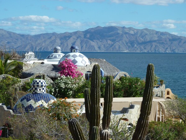 A coastal landscape with thatched-roof structures, cacti, and a view of the sea and mountains.