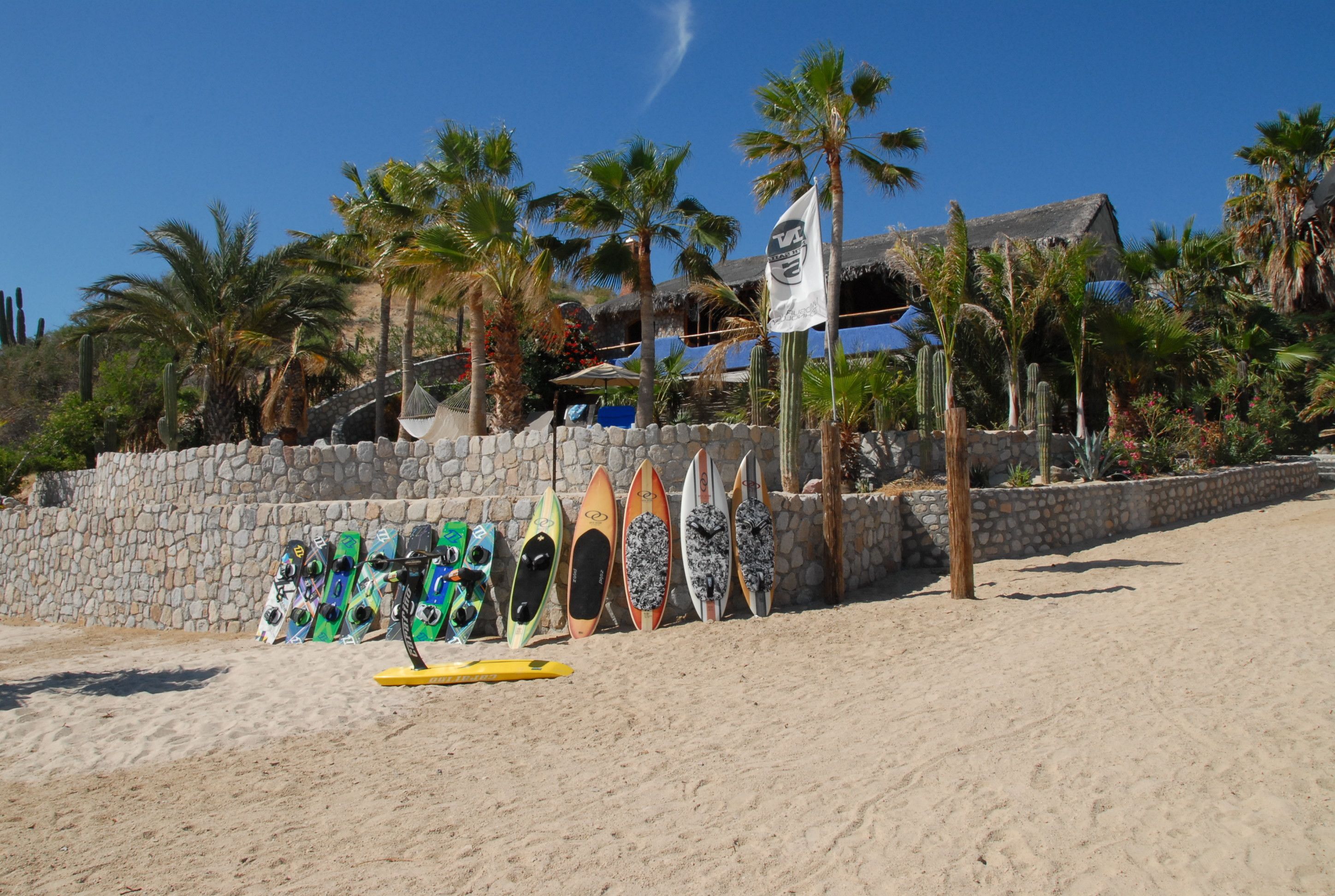 Ein sandiger Strand mit mehreren Surfbrettern, die an eine Steinmauer gelehnt sind, Palmen und ein Gebäude mit einem Strohdach im Hintergrund.