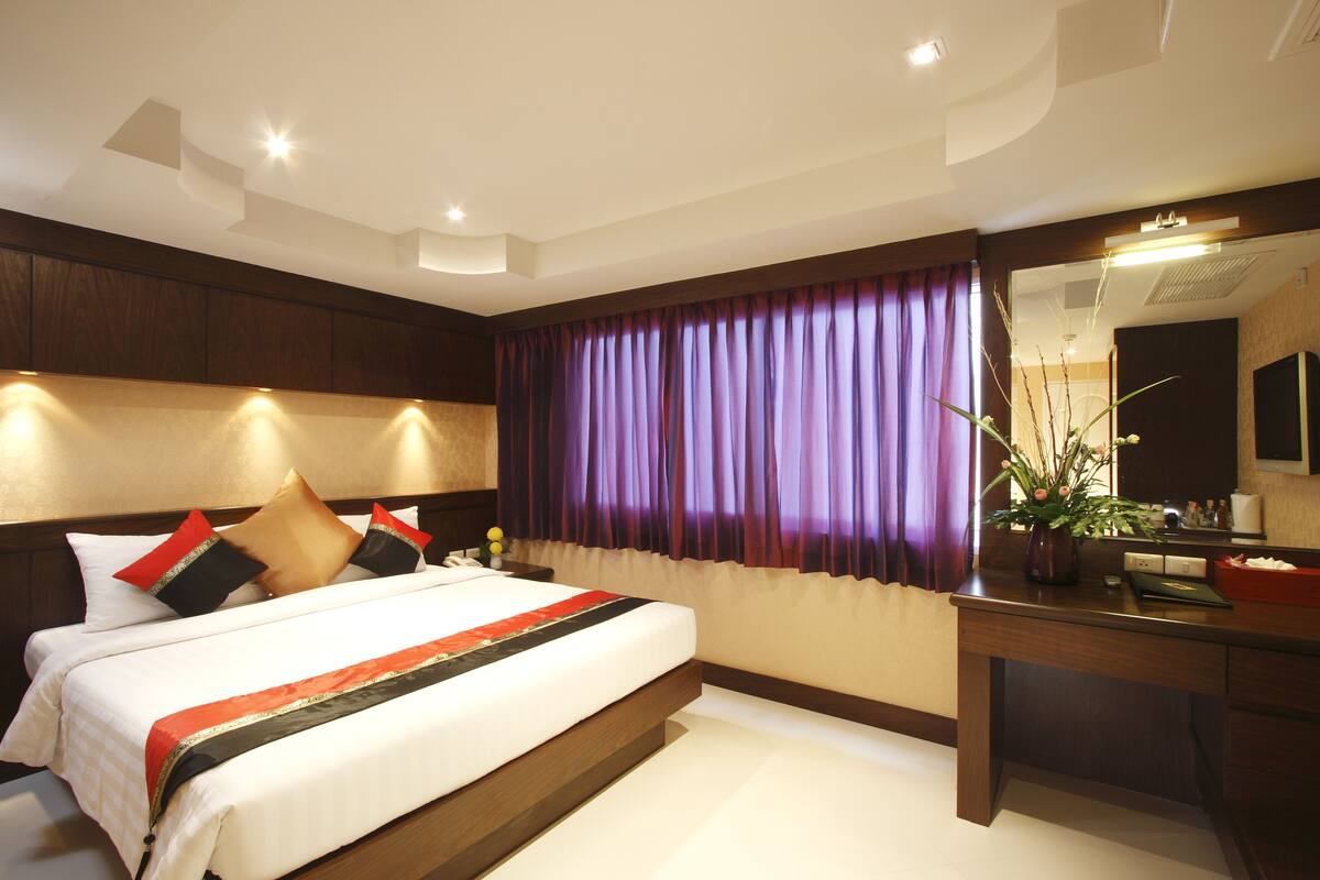 A modern hotel room with a large bed, a wooden headboard, a desk with a mirror, and a window with purple curtains.
