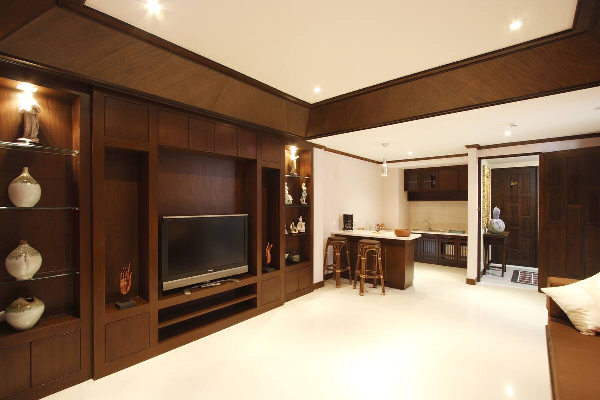 A modern living room with a flat-screen TV, wooden shelving, and a kitchen area in the background.
