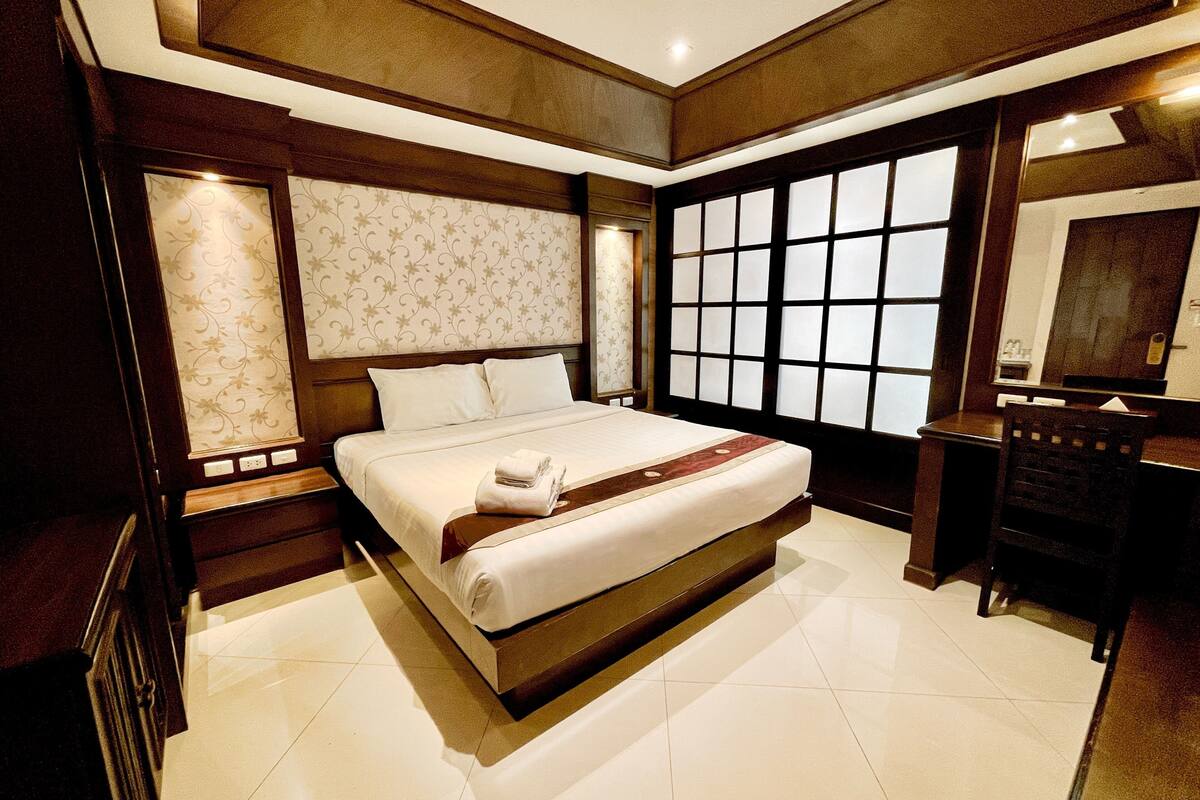 A hotel room with a large bed, a wooden headboard, a large window with blinds, a desk with a chair, and a mirror.