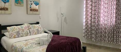 Un dormitorio con cama, mesita de noche, lámpara y ventana con cortinas estampadas.