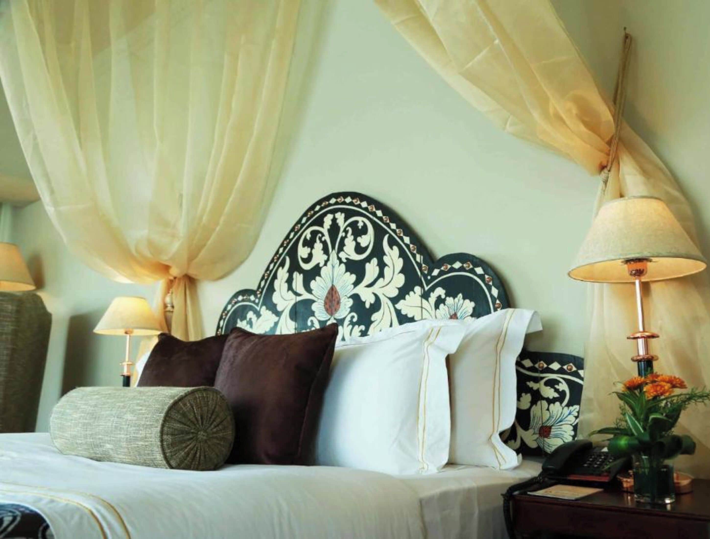 Una cama bien hecha con un cabecero decorativo, dos lámparas de noche, una mesita con un teléfono y un jarrón con flores, junto a cortinas translúcidas.