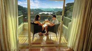 A couple sitting on wicker chairs on a balcony with a glass door, overlooking a scenic view of a lake and mountains. - Thilanka Hotel (Kandy)