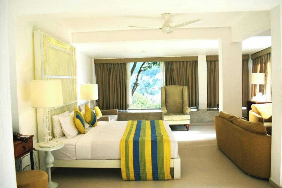 A hotel room with a bed, a sofa, a chair, and a desk. There is a window with curtains, a ceiling fan, and a lamp on the desk.