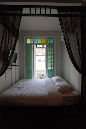 A bedroom with a bed, curtains, and a view of a balcony through a window with green shutters. - 99 OLDTOWN BOUTIQUE GUESTHOUSE (Phuket)