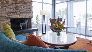 Un salon moderne avec une cheminée en pierre, un canapé bleu agrémenté de coussins orange, une table ronde sur laquelle est posé un vase de fleurs, et de grandes fenêtres offrant une vue sur l’extérieur.