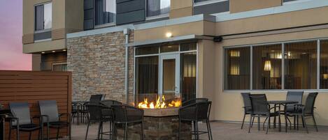 A modern outdoor seating area with a stone fire feature surrounded by chairs, set against a building with multiple windows and a stone wall.
