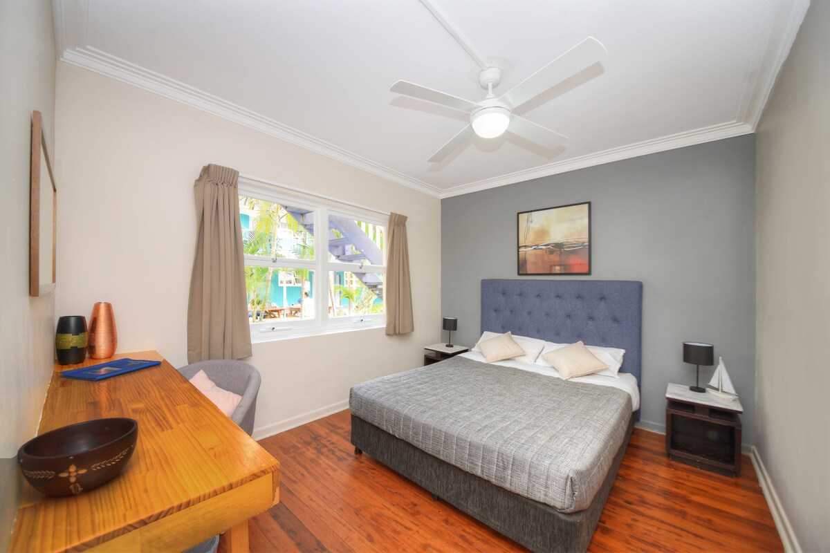 A bedroom with a bed, a desk, a chair, a window with curtains, a ceiling fan, and a painting on the wall.