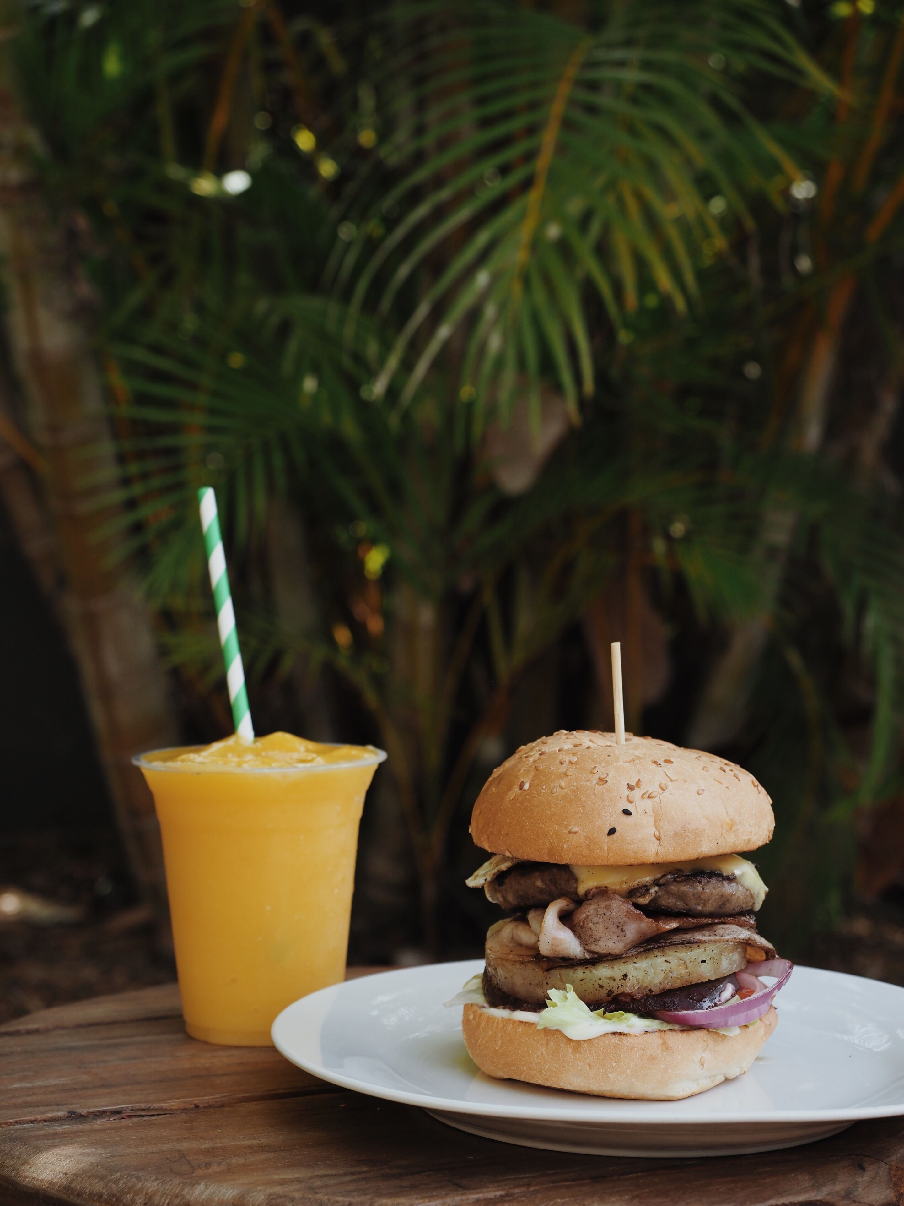 A burger with a toothpick on a white plate, a yellow drink with a striped straw, and a wooden table.