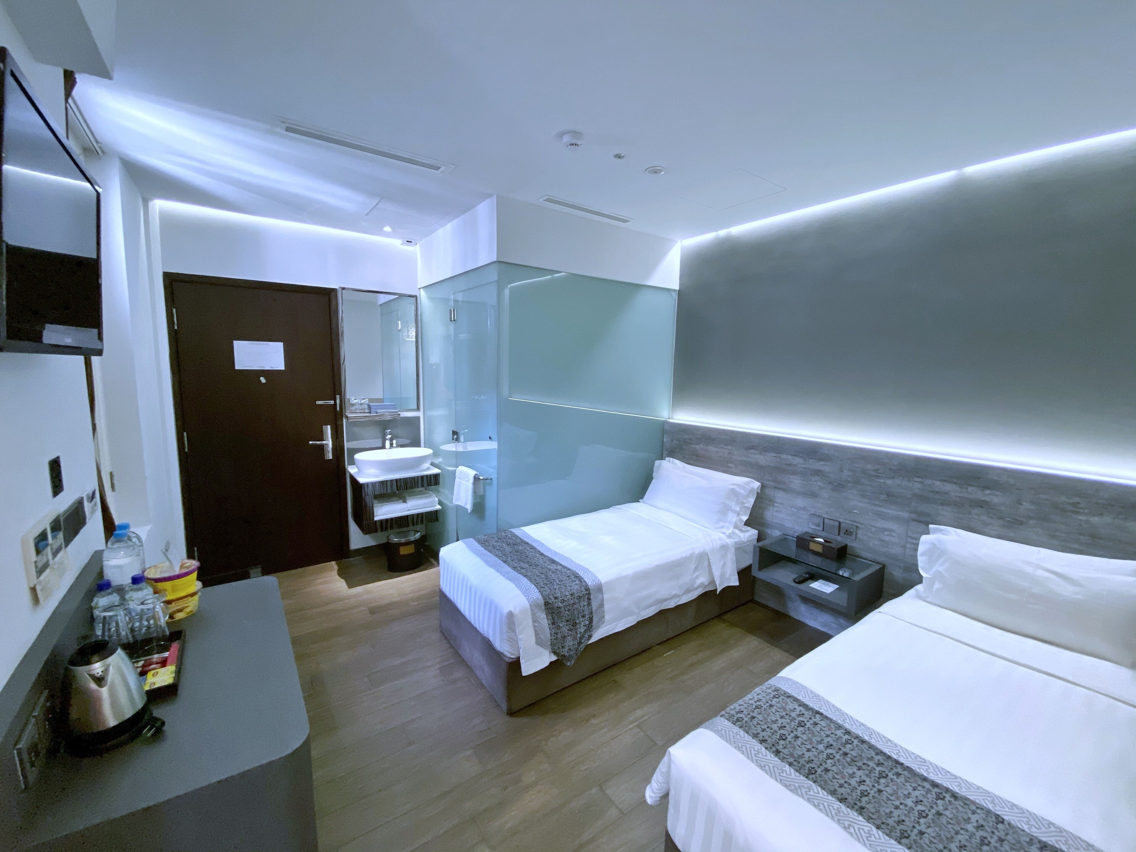 Une chambre d’hôtel moderne dotée d’un grand lit, d’un lit plus petit, d’un bureau avec une bouilloire, d’un miroir et d’une salle de bain avec une douche à parois de verre.