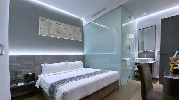 Una habitación de hotel moderna con una cama grande, un mamparo de vidrio, un baño con lavamanos y espejo, y una pequeña zona de estar.