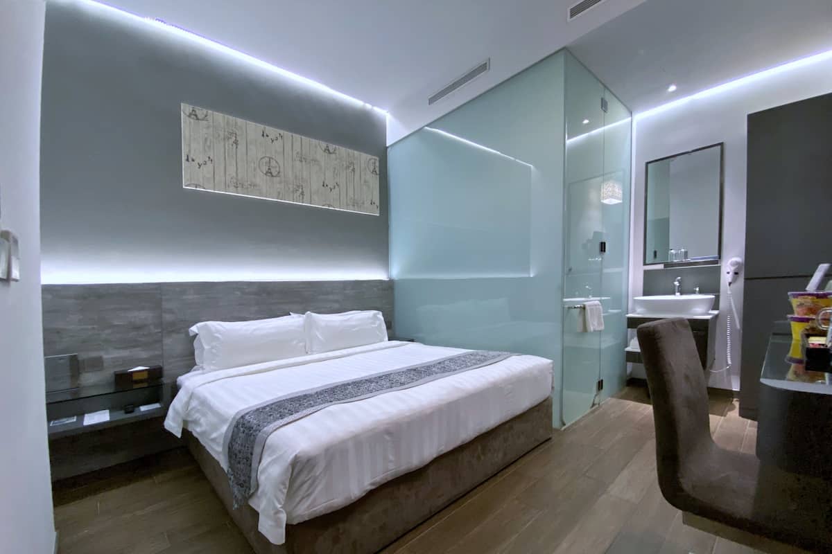 Une chambre d’hôtel moderne dotée d’un grand lit, d’une cloison en verre, d’une salle de bain avec un lavabo et un miroir, et d’un petit coin salon.