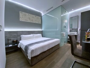 Una habitación de hotel moderna con una cama grande, un mamparo de vidrio, un baño con lavamanos y espejo, y una pequeña zona de estar.