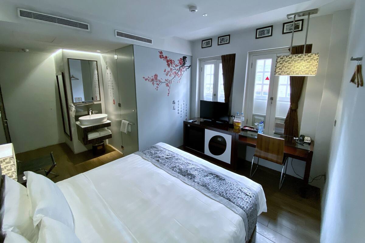 Une chambre d’hôtel comprenant un lit, un bureau avec un ordinateur, une télévision, une salle de bain avec un lavabo et un miroir, et une fenêtre avec des rideaux.