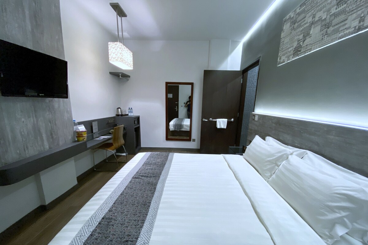 Una habitación de hotel moderna con una cama grande, un escritorio, una silla, un televisor de pantalla plana en la pared y un espejo.