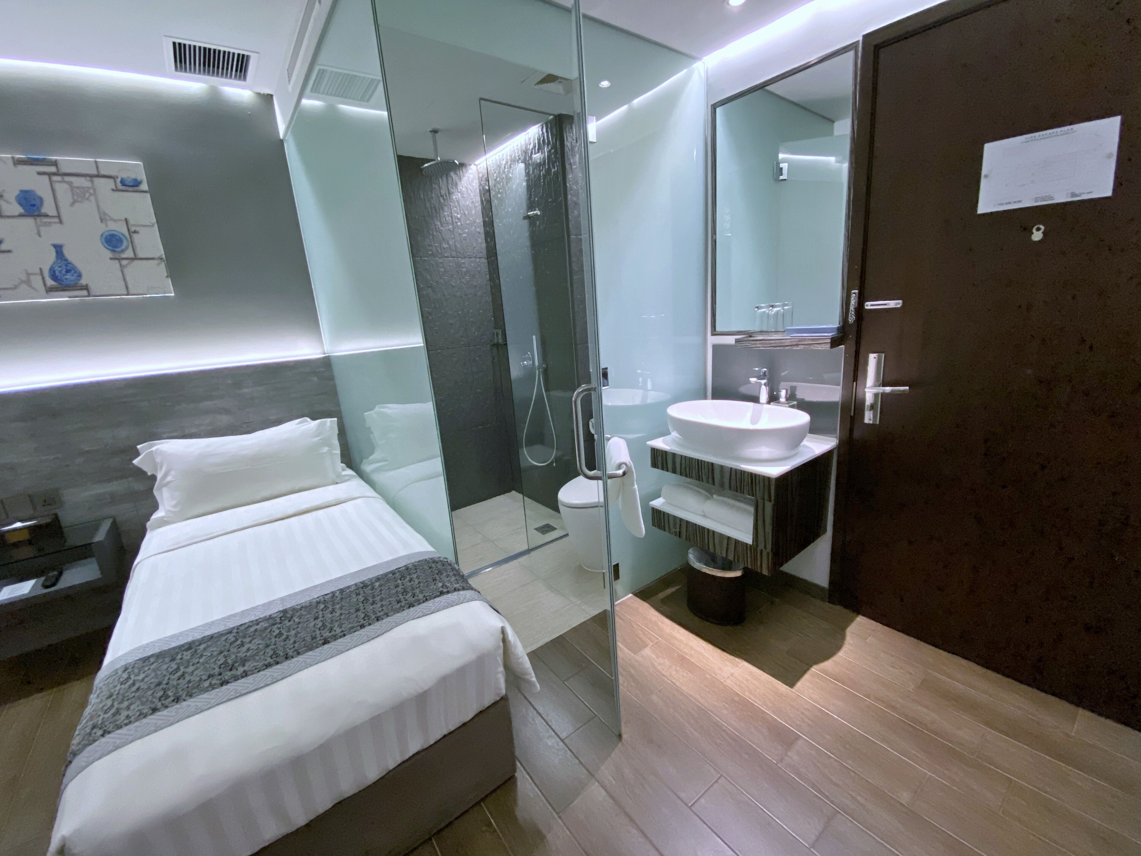 A modern hotel room with a bed, a glass-enclosed shower, a sink, and a mirror.