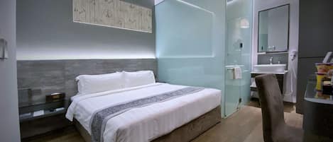 Quarto de hotel moderno com uma cama grande, divisória de vidro, banheiro com pia e uma cadeira.