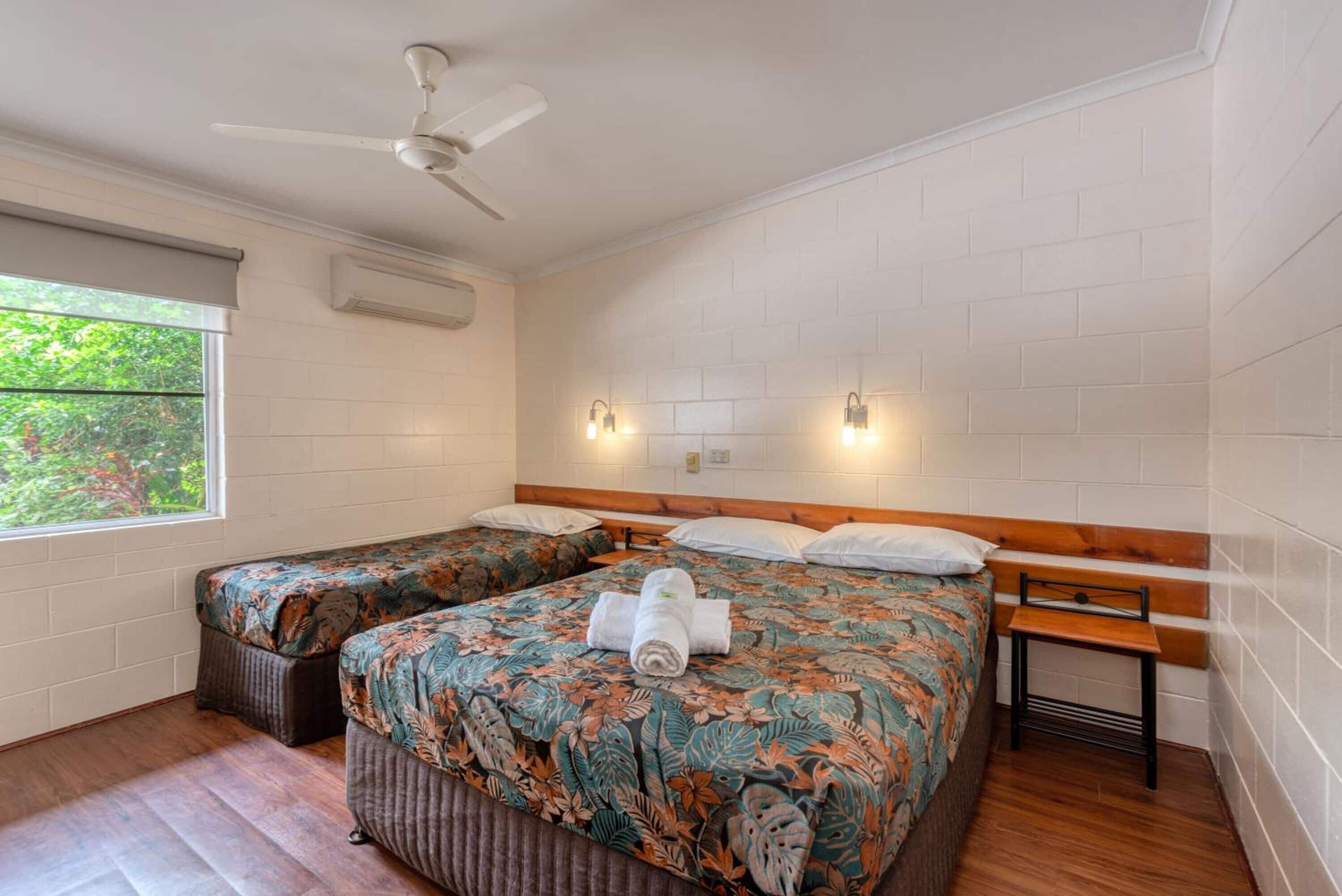 A room with two beds, a ceiling fan, a window with a view of greenery, and a wall-mounted air conditioner.