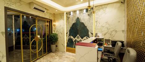 A reception area with marble flooring, a reception desk, two chairs, a potted plant, and a decorative wall mural.