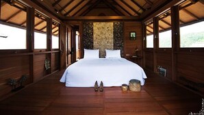 A large, well-lit bedroom with a wooden ceiling and walls, a large bed, and a view of the outdoors through large windows. - Sanak Retreat Bali (Gobleg)