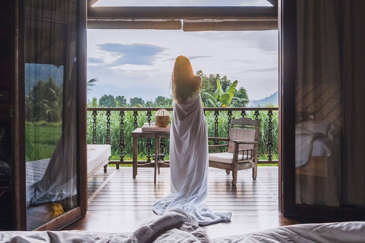 A room with a bed, a table, and a chair on a balcony with a view of greenery.