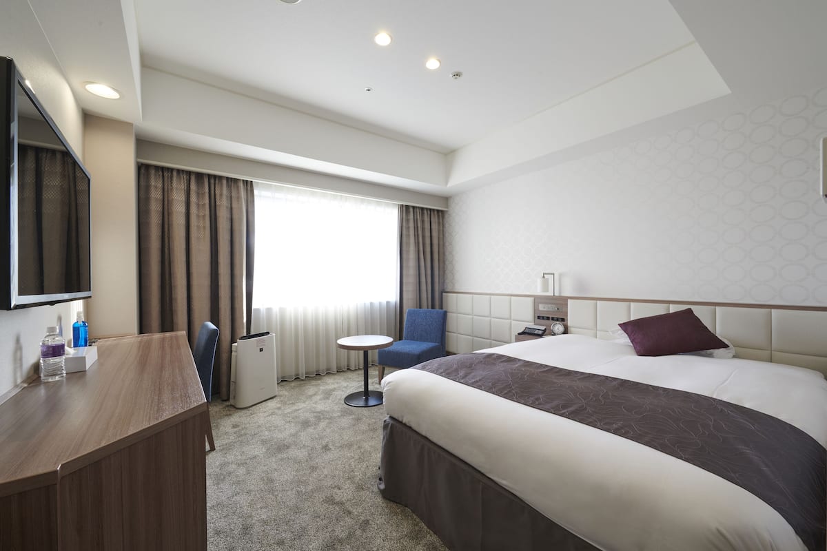 A hotel room with a bed, a desk, a chair, a small table, and a window with curtains.