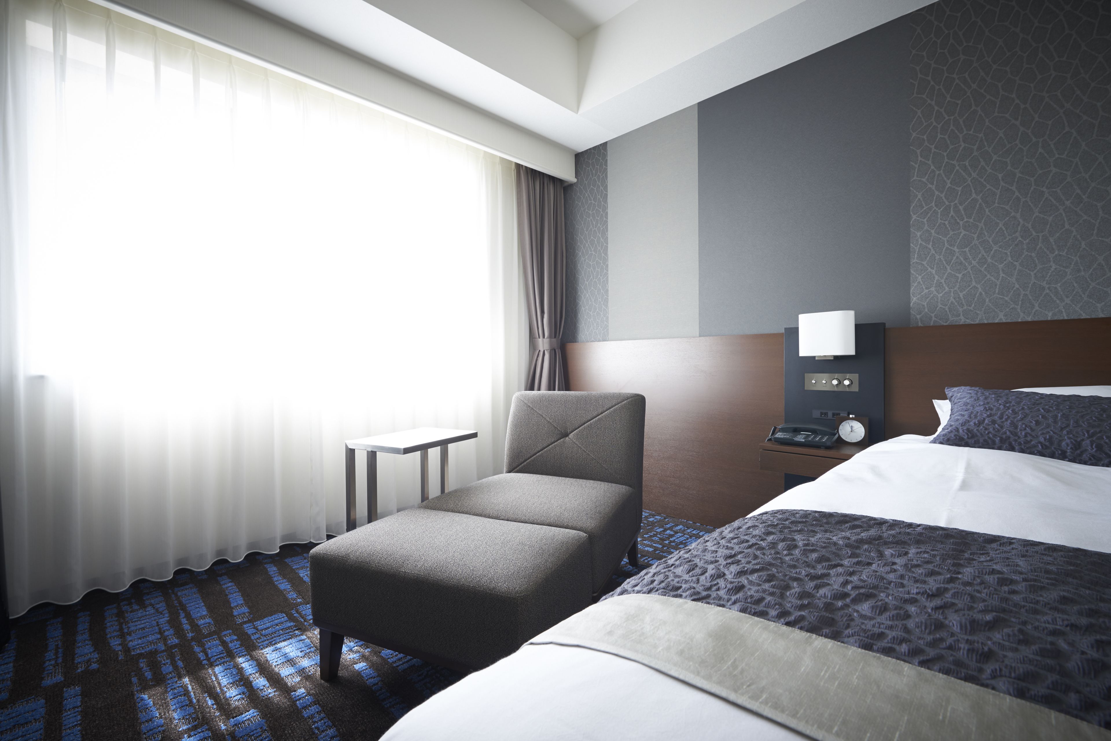 Une chambre d’hôtel avec un lit, une chaise, une table de chevet, une lampe et une fenêtre avec des rideaux.