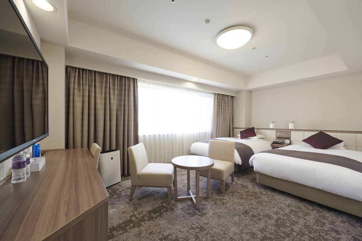 A hotel room with a bed, two chairs, a small table, a desk, a TV, and a window with curtains.