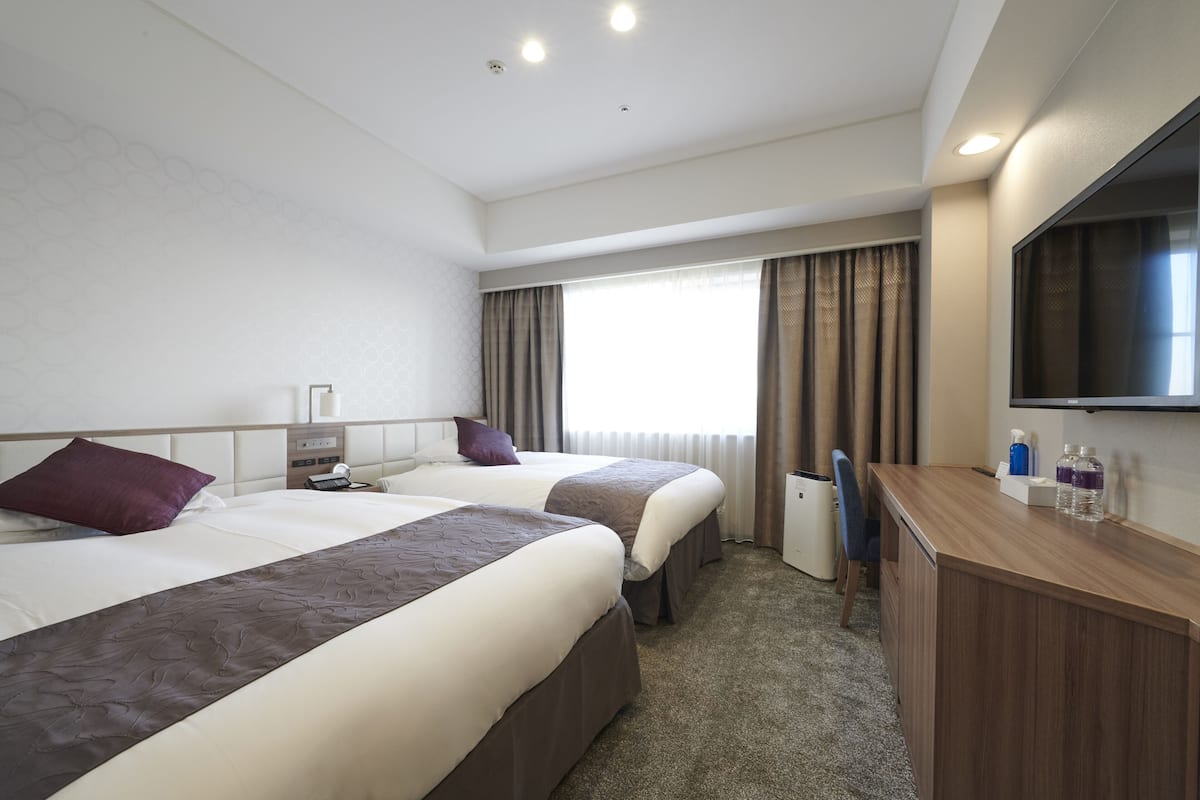 A hotel room with two beds, a desk, a TV, and a window with curtains.