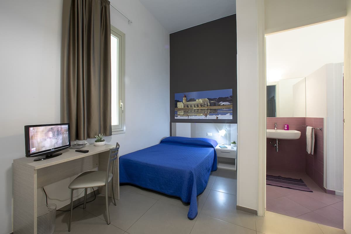 A hotel room with a desk, two beds, a TV, a window with curtains, a painting, and a bathroom.