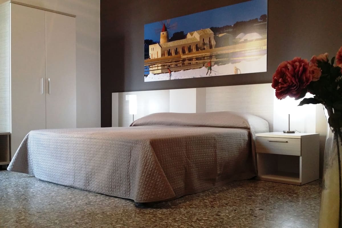 A bedroom with a bed, a nightstand, a vase with flowers, and a painting of a building on the wall.