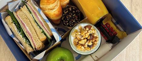 Una caja de comida con sándwiches, un croissant, una manzana, un vaso de jugo de naranja, una galleta y una barra de granola.