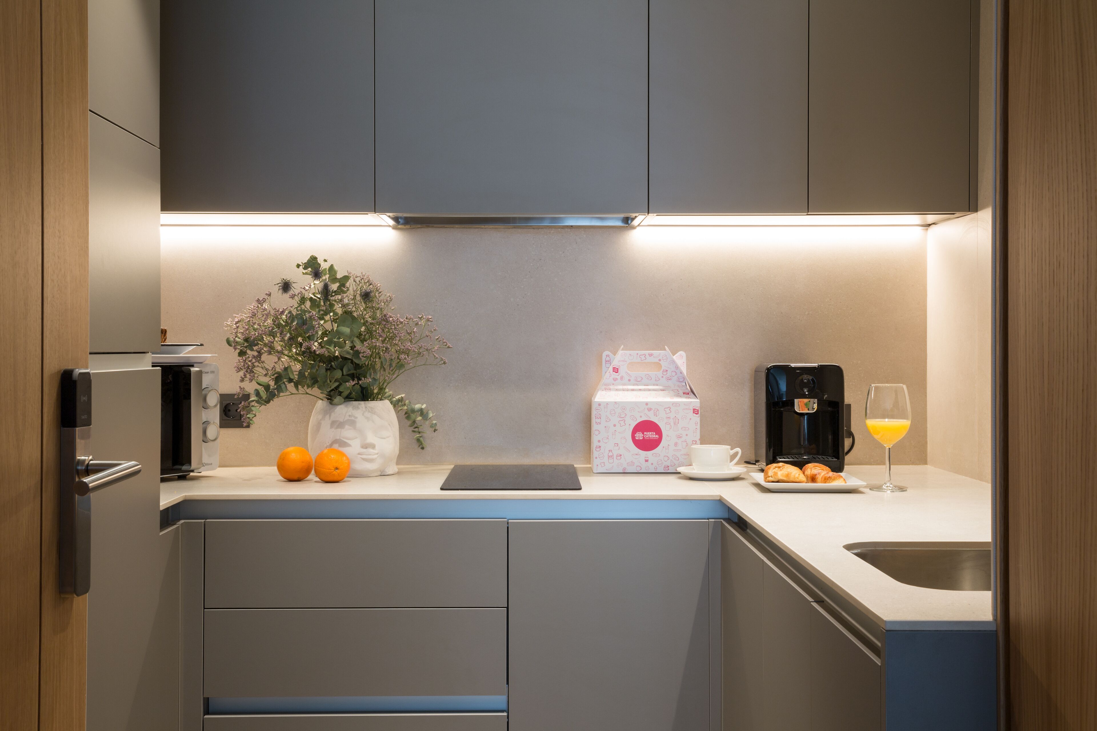 Una cocina moderna con cafetera integrada, un lavamanos y una encimera con planta en maceta y naranjas.