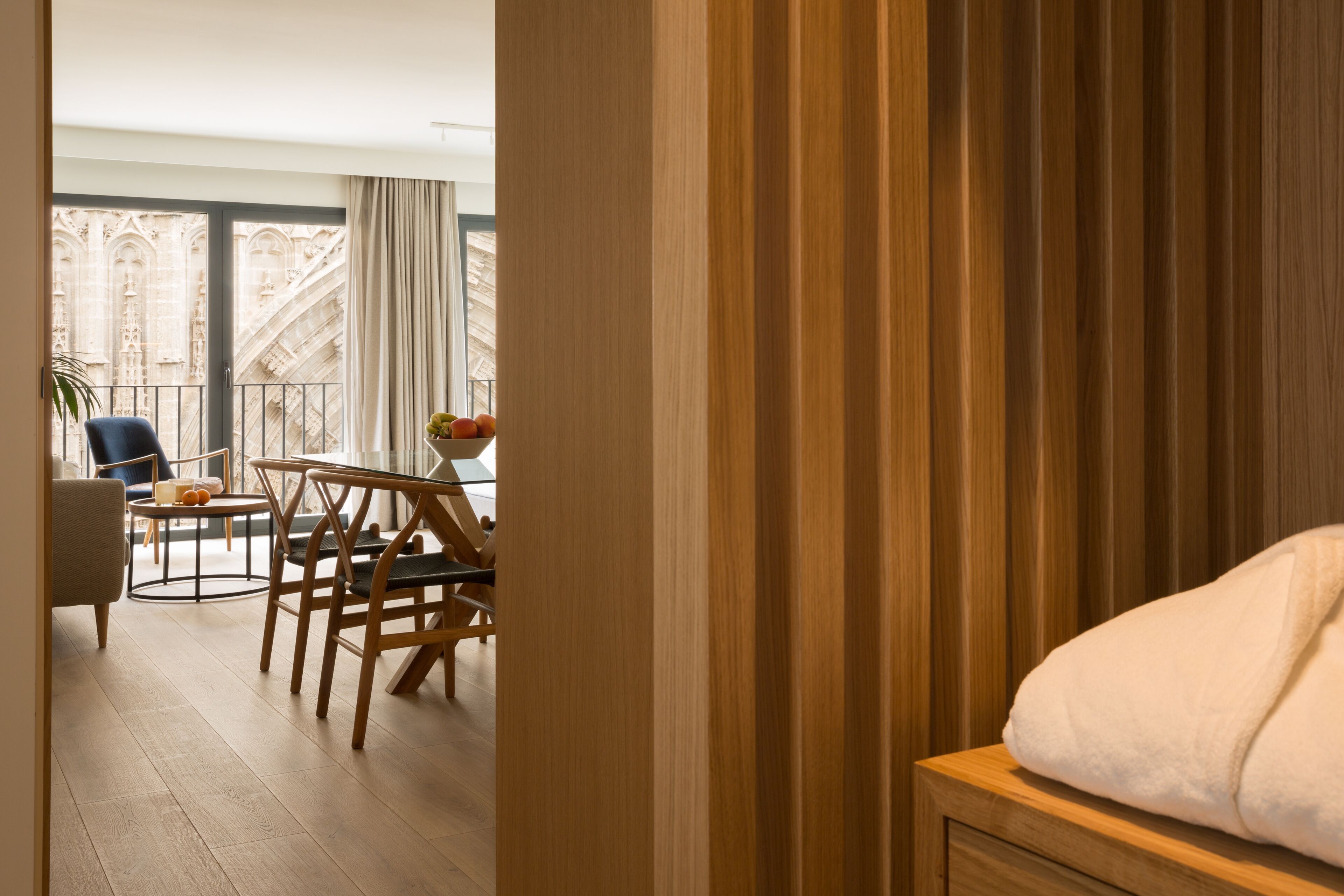 Habitación de hotel con suelo de madera, cama con manta blanca, mesa de madera con sillas y vistas a una catedral a través de amplios ventanales.