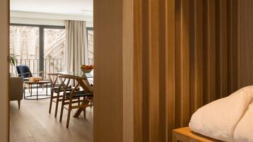 Habitación de hotel con suelo de madera, cama con manta blanca, mesa de madera con sillas y vistas a una catedral a través de amplios ventanales.