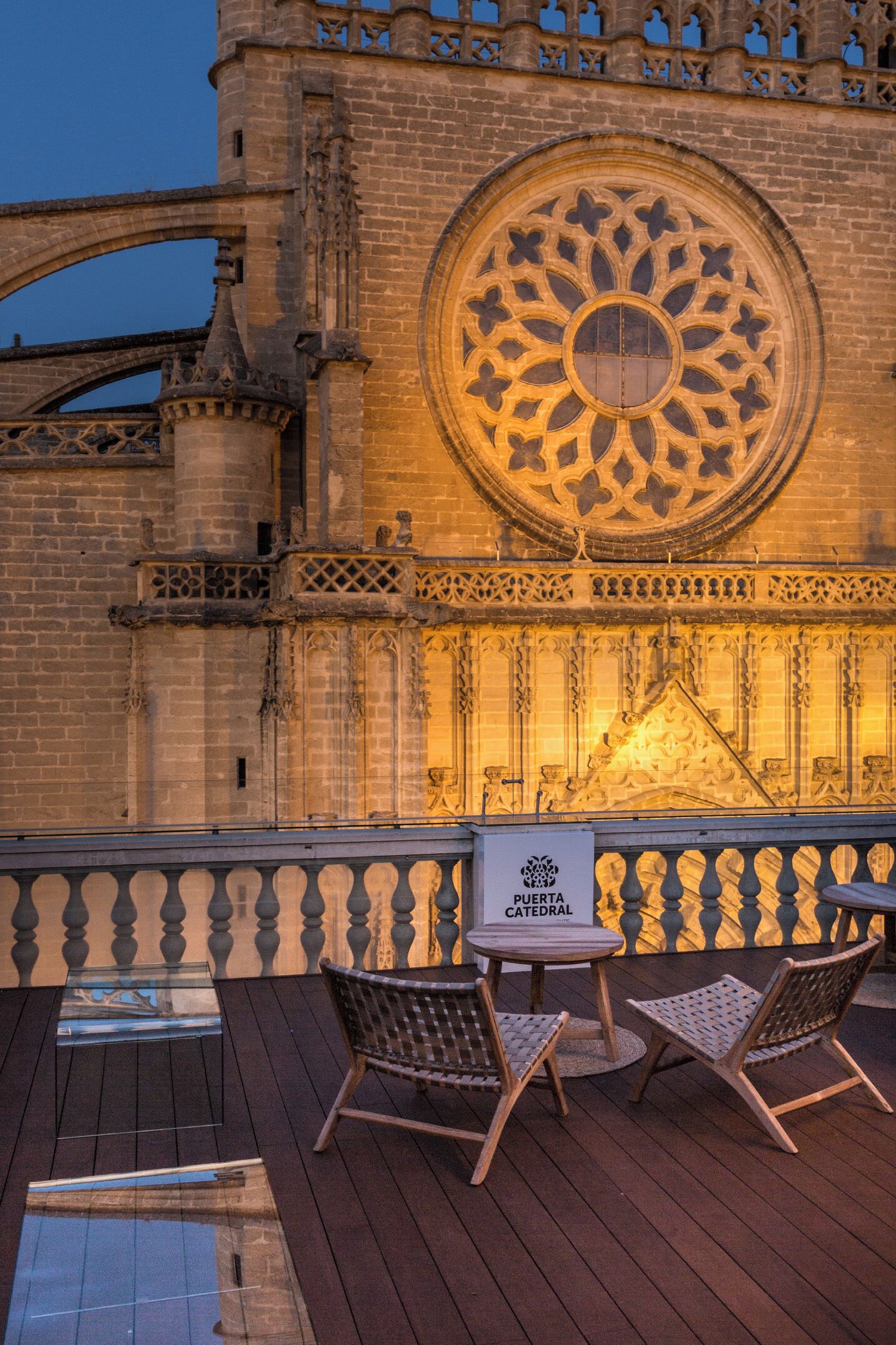 A view of Puerta Catedral with a large ornate window and a wooden deck with chairs.