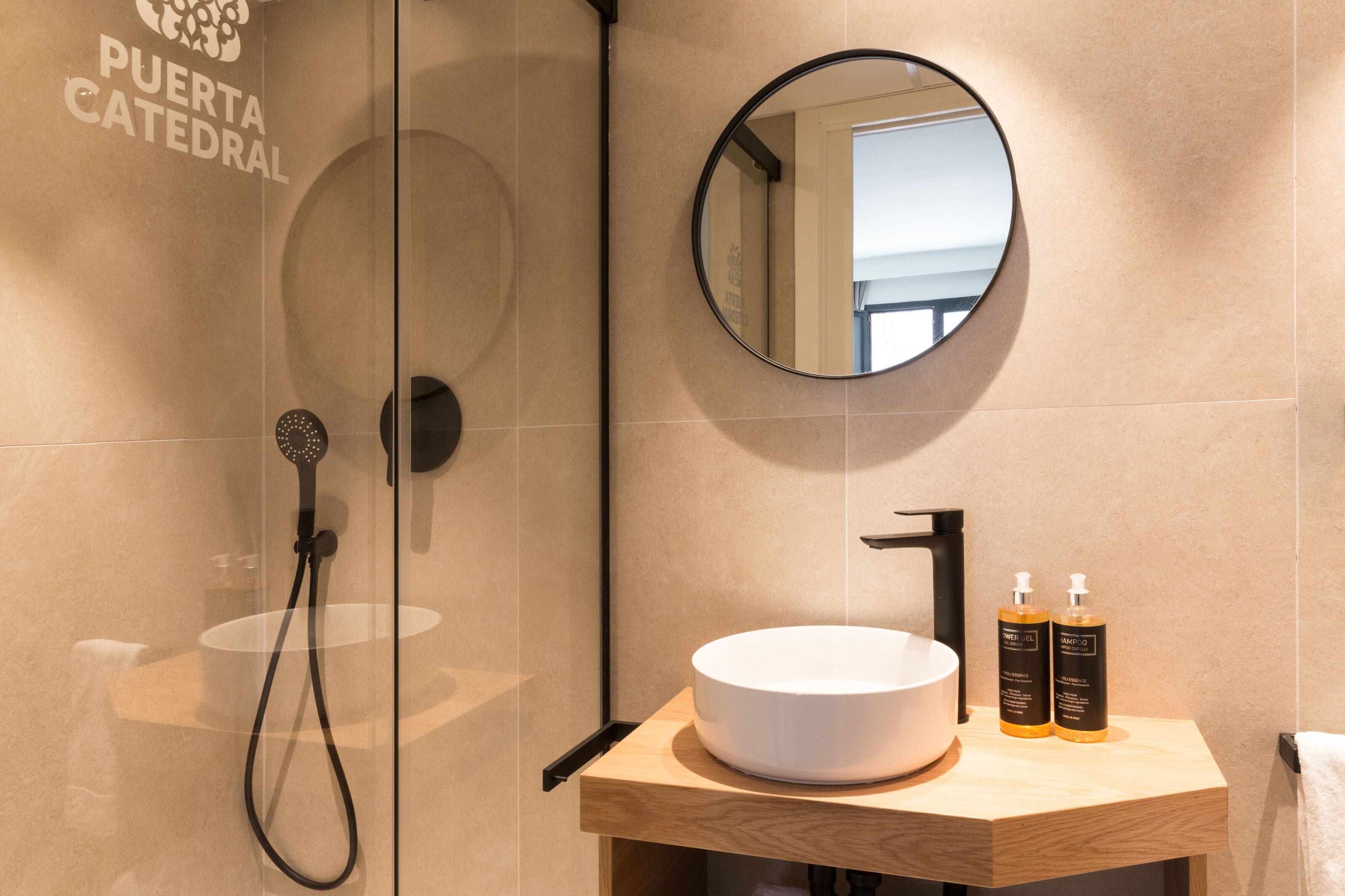 Un baño moderno con cabina de ducha de vidrio, un espejo redondo, tocador de madera con lavabo blanco y dos botellas de champú.
