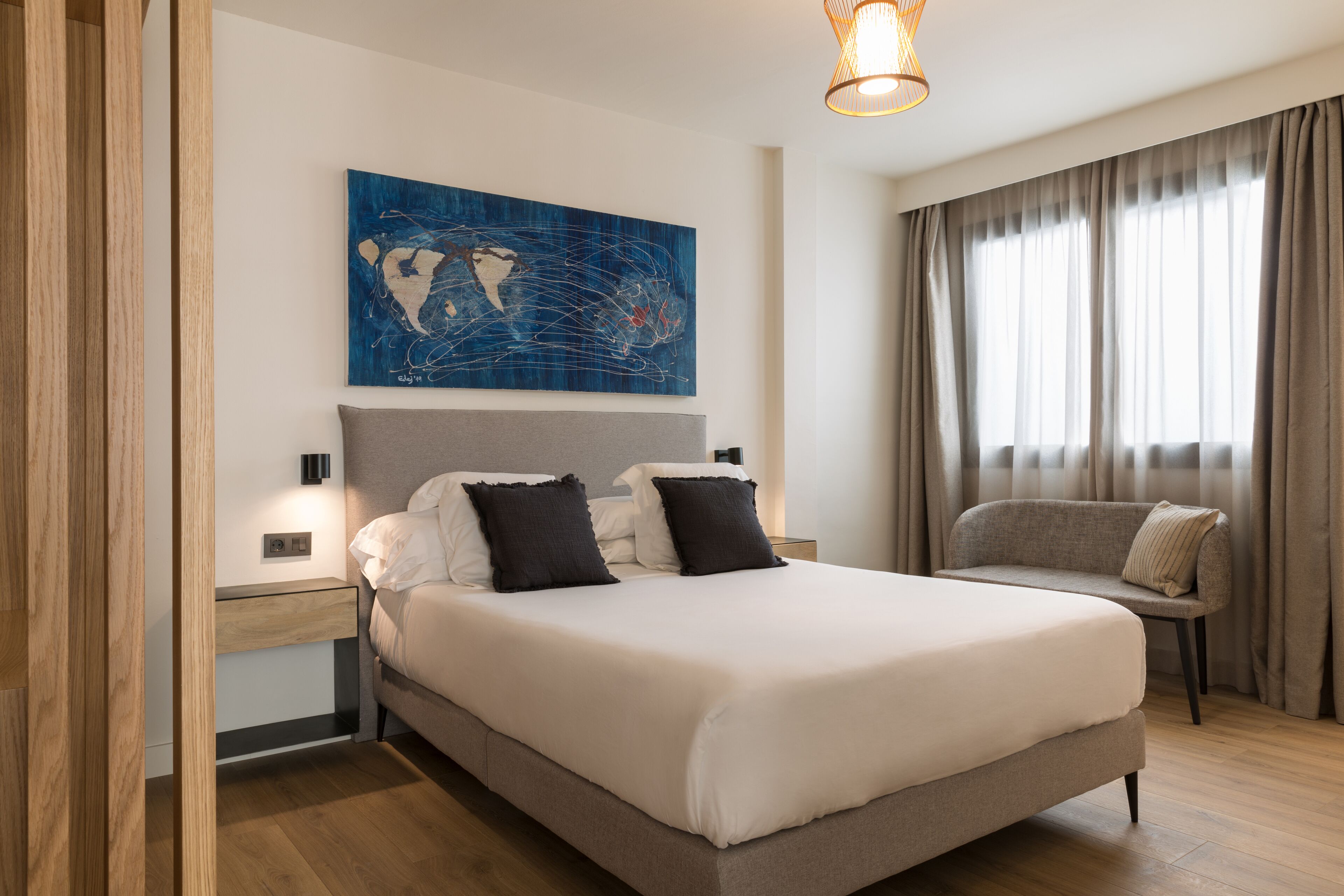 A modern hotel room with a large bed, a bedside table, a nightstand, a sofa, and a large window with curtains.