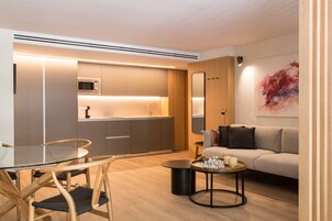A modern living space with a glass dining table, wooden chairs, a round coffee table, a sofa, and a kitchen area with built-in appliances.