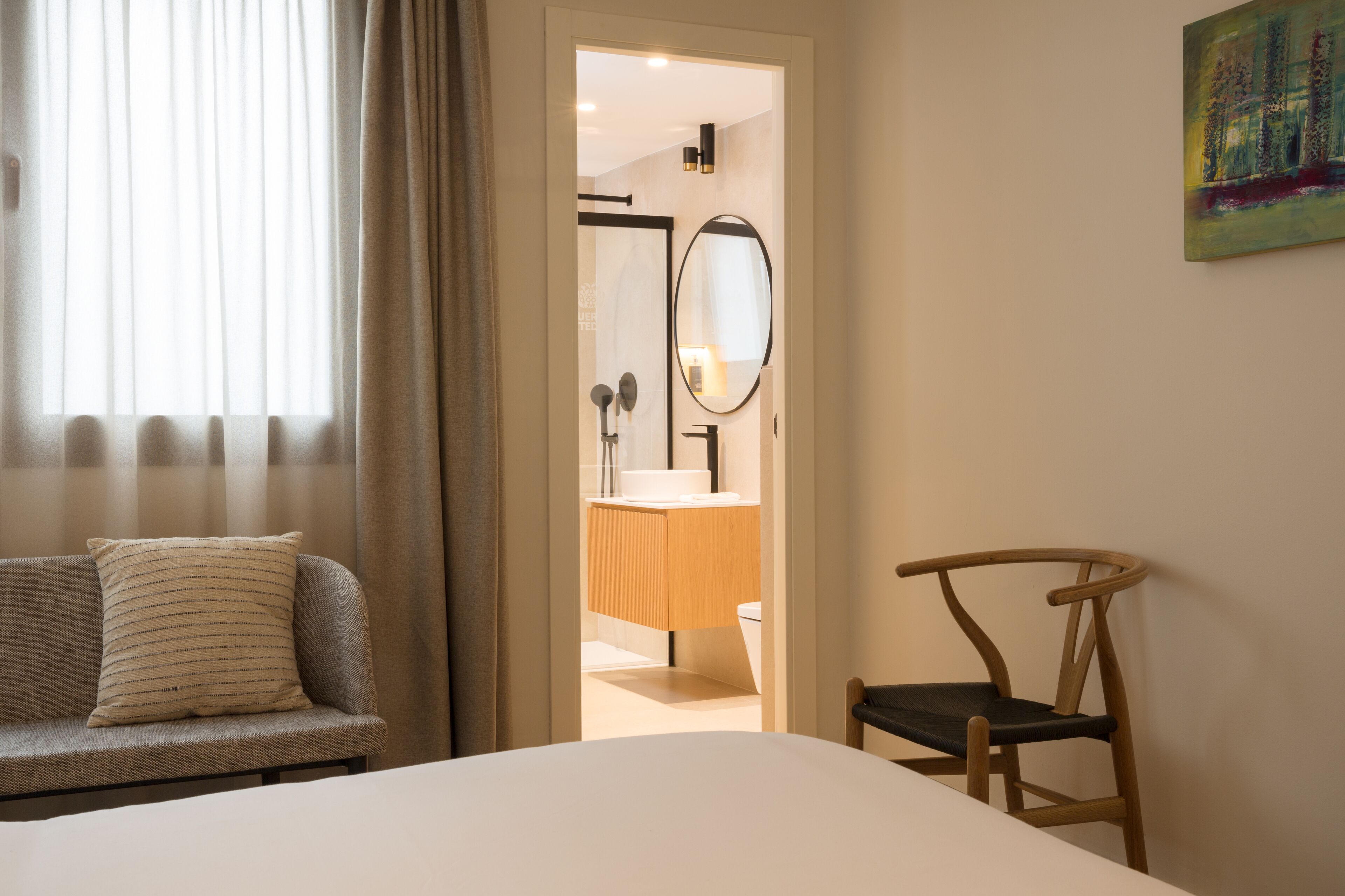 A bedroom with a bed, a wooden chair, a sofa with a striped pillow, and a bathroom visible through an open door.