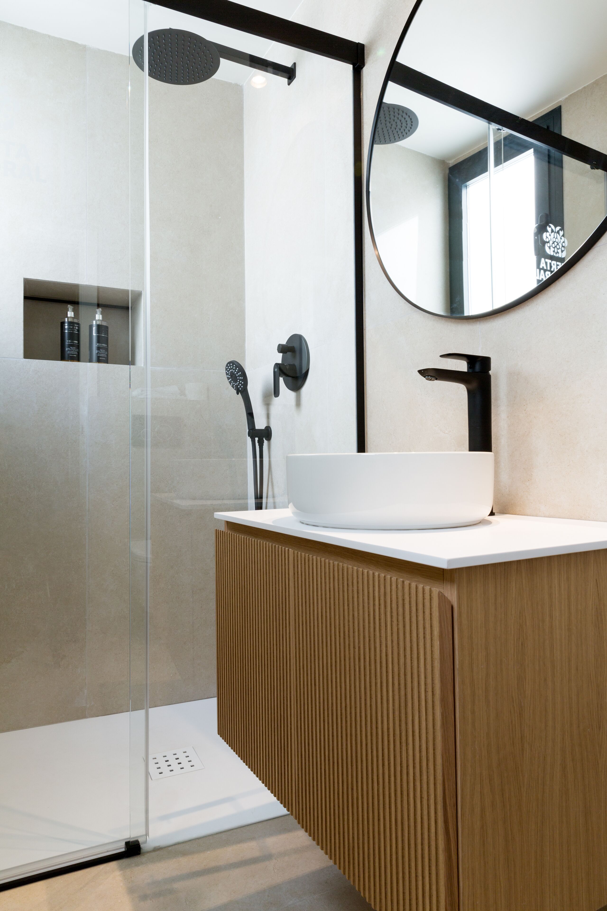 A modern bathroom with a glass shower enclosure, a wooden vanity, a white sink, and a round mirror.