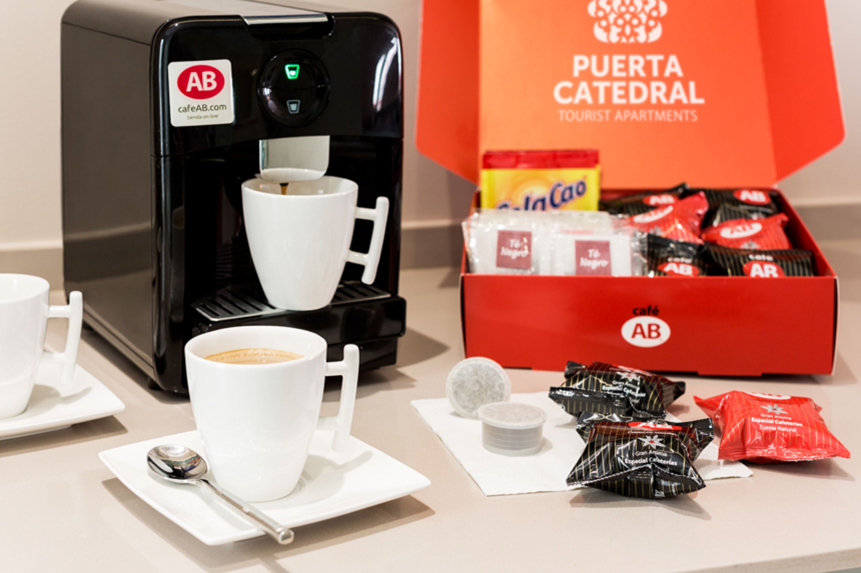 A coffee machine with two cups, a spoon, and a box of AB brand snacks.