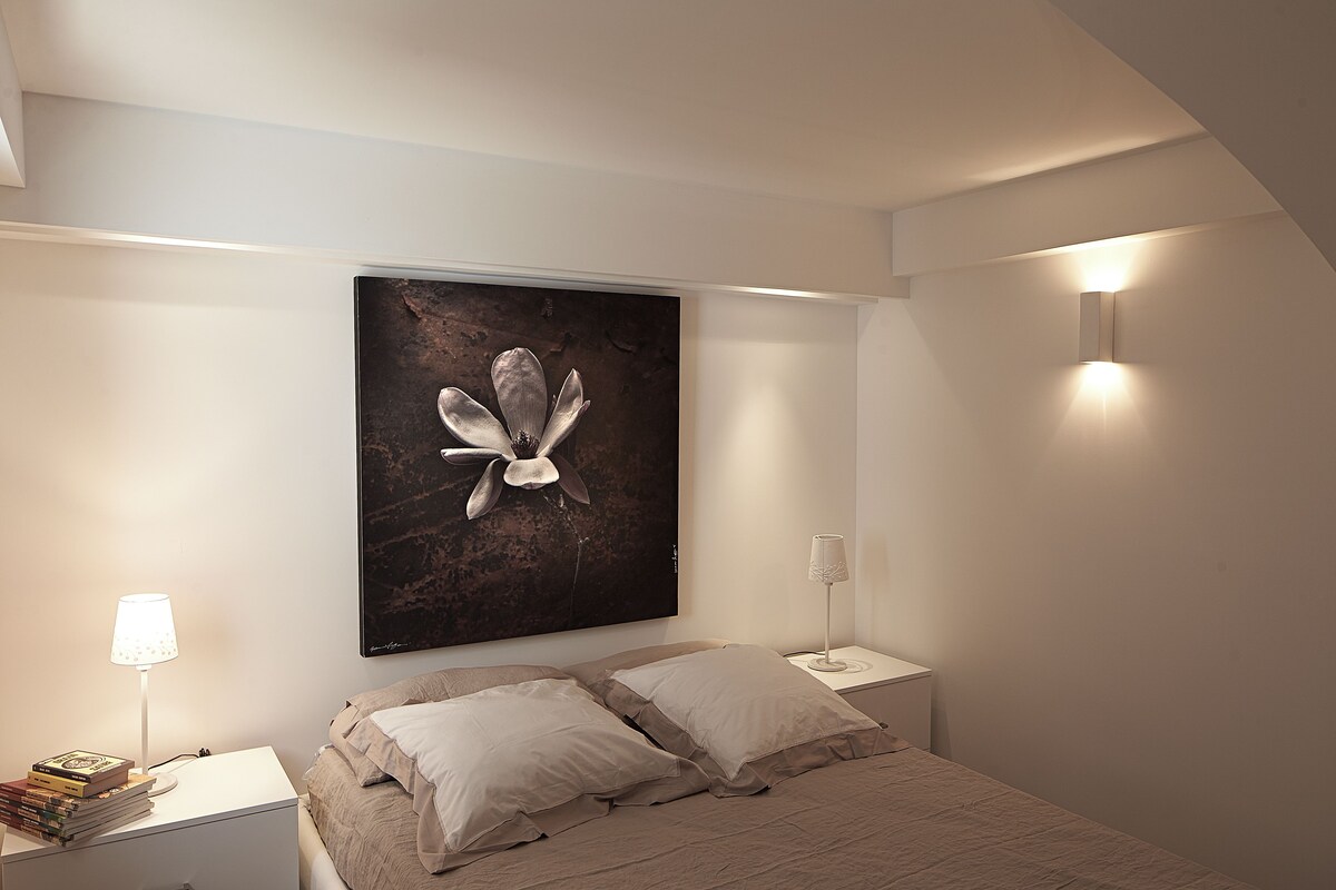 A bedroom with a bed, two bedside tables, a wall-mounted lamp, a framed artwork, and a floor lamp.