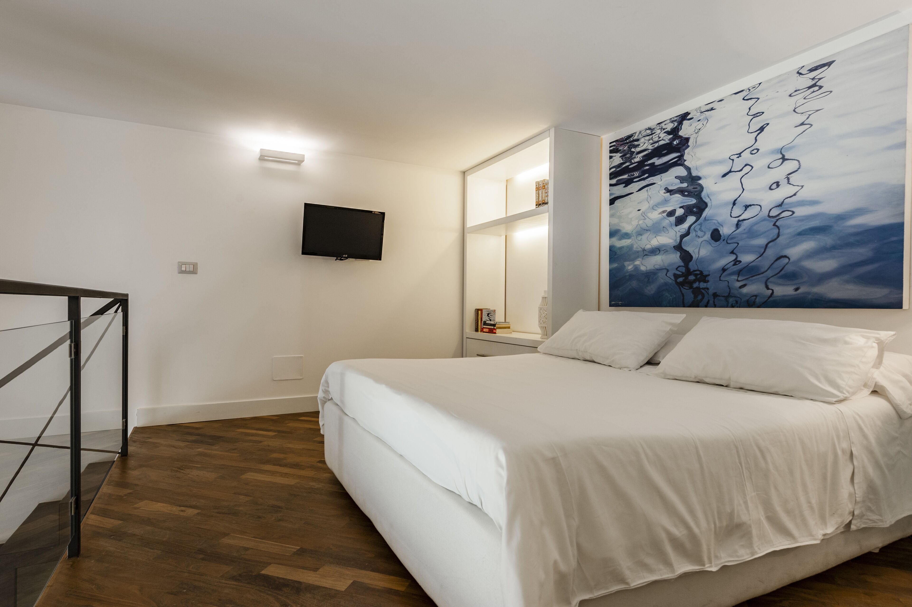 Un dormitorio con piso de madera, una cama con ropa de cama blanca, un televisor montado y un cuadro abstracto grande en la pared.