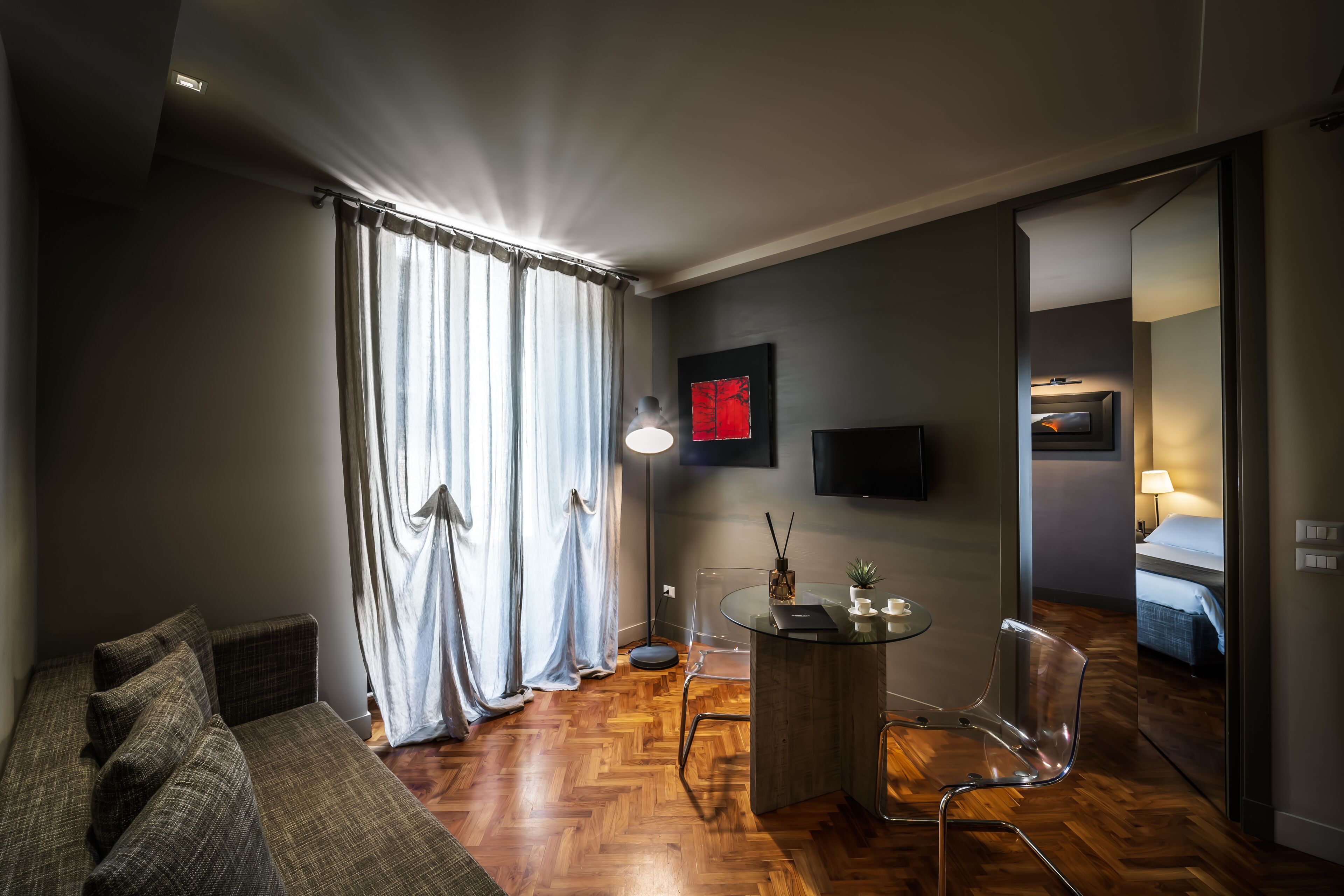 A modern hotel room with a wooden floor, a sofa, a dining table with chairs, a TV mounted on the wall, and a bed in the adjacent room.