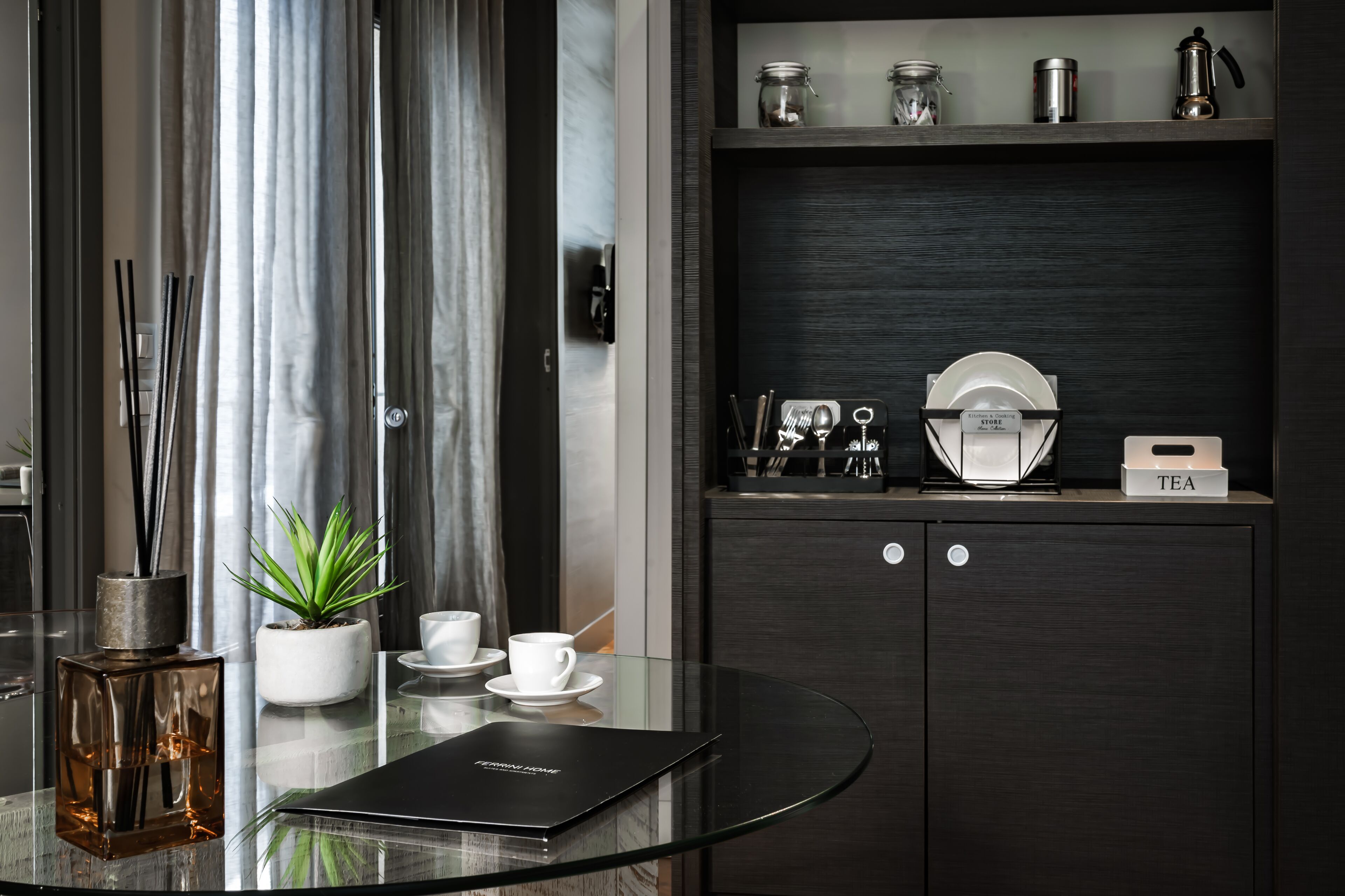 A modern room with a glass table, a potted plant, a bottle of perfume, and a black book. There is a dark wooden cabinet with kitchen items and a tea station.