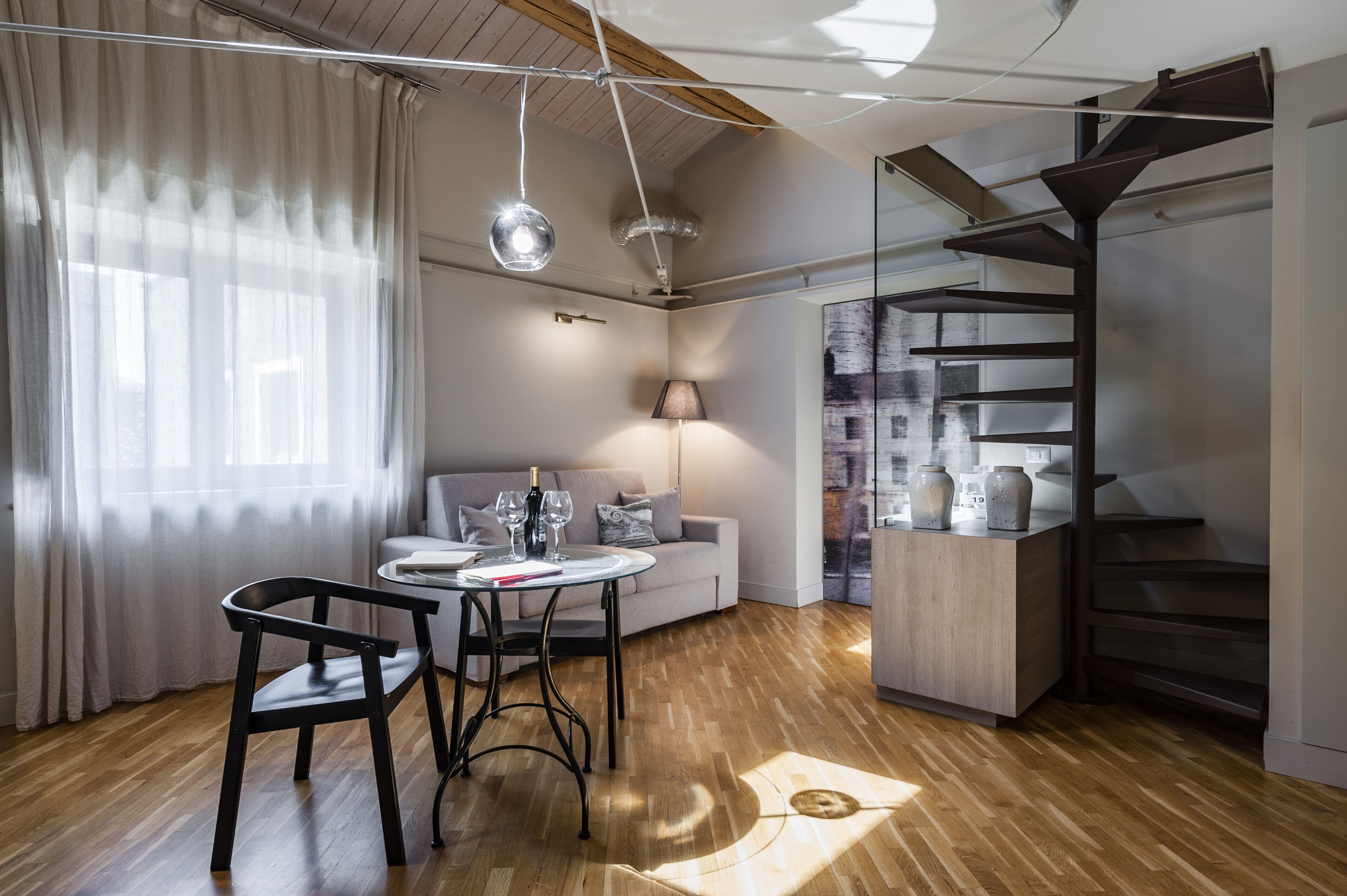 A modern living space with a wooden floor, a round table with chairs, a sofa, a lamp, and a spiral staircase.