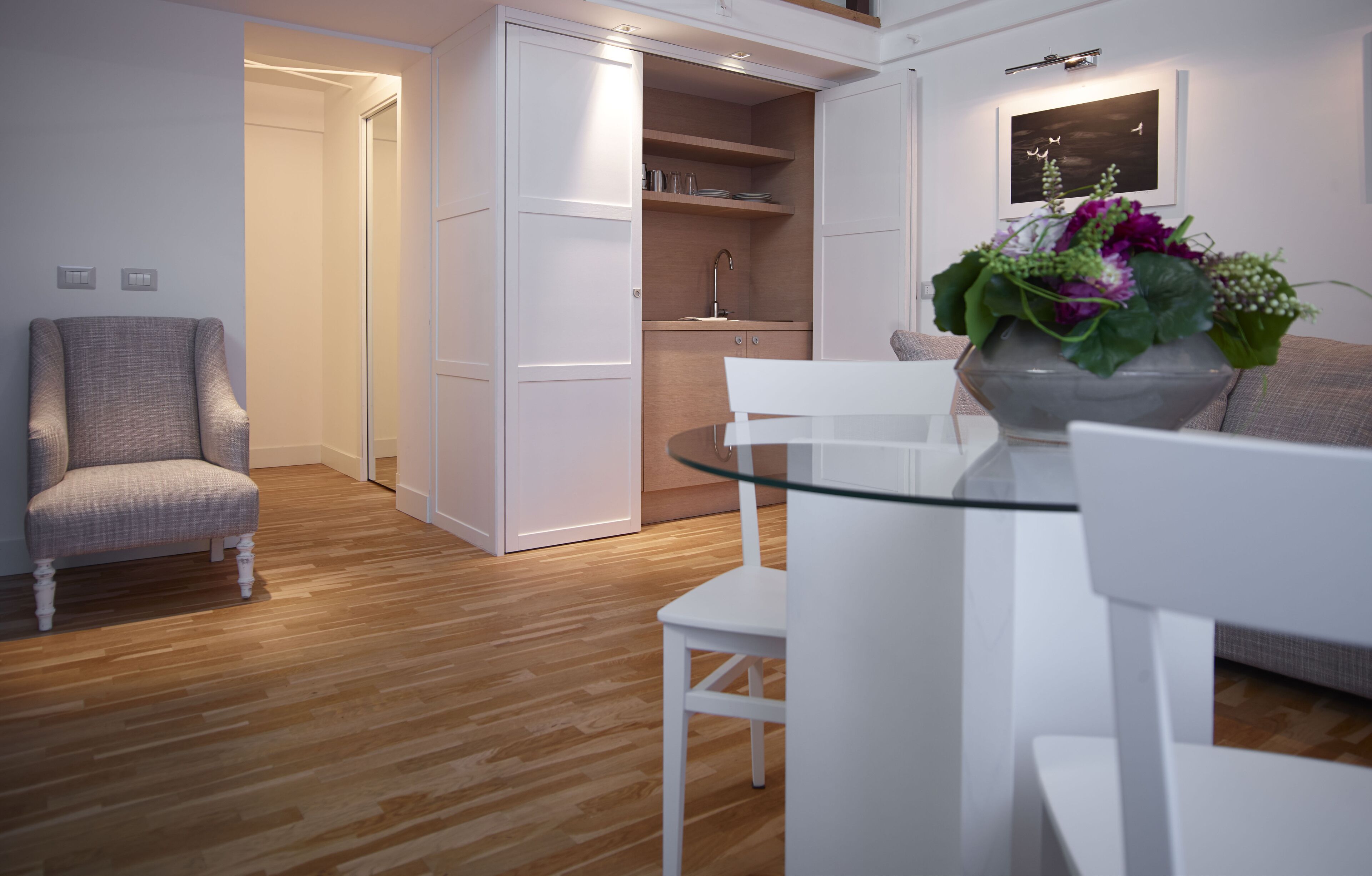 Ein modernes Wohnzimmer mit einem Esstisch mit Glasplatte, einem weißen Sofa, einem grauen Sessel und einer integrierten Küchenzeile mit Holzschränken und einer Spüle.